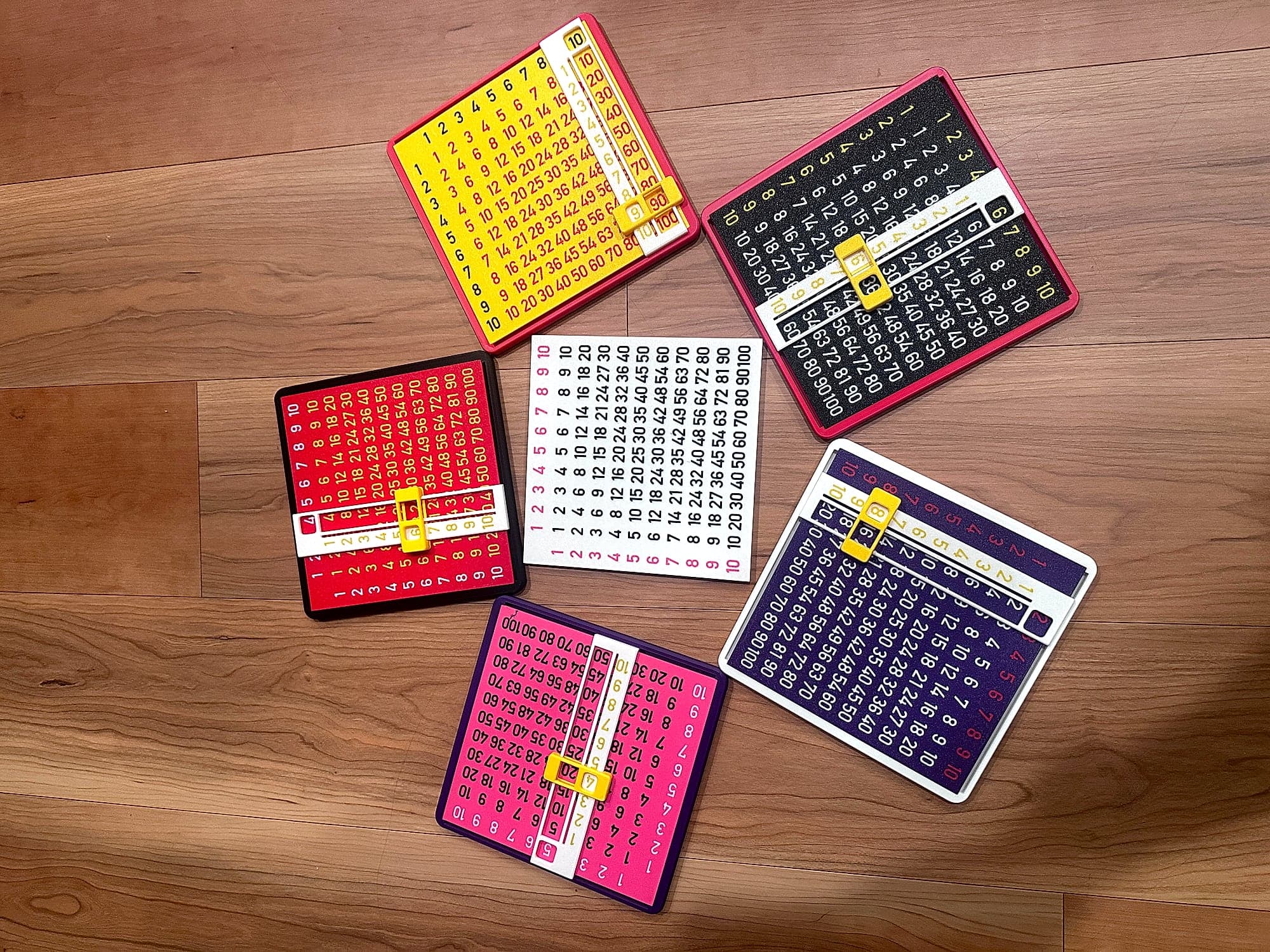 Six multiplication slider boards in different colorways arranged on a wood floor